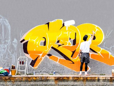 Street artist painting colored graffiti on public space wall - Modern art concept of urban guy performing and preparing live murales paint with yellow aerosol color spray - Cloudy afternoon filter